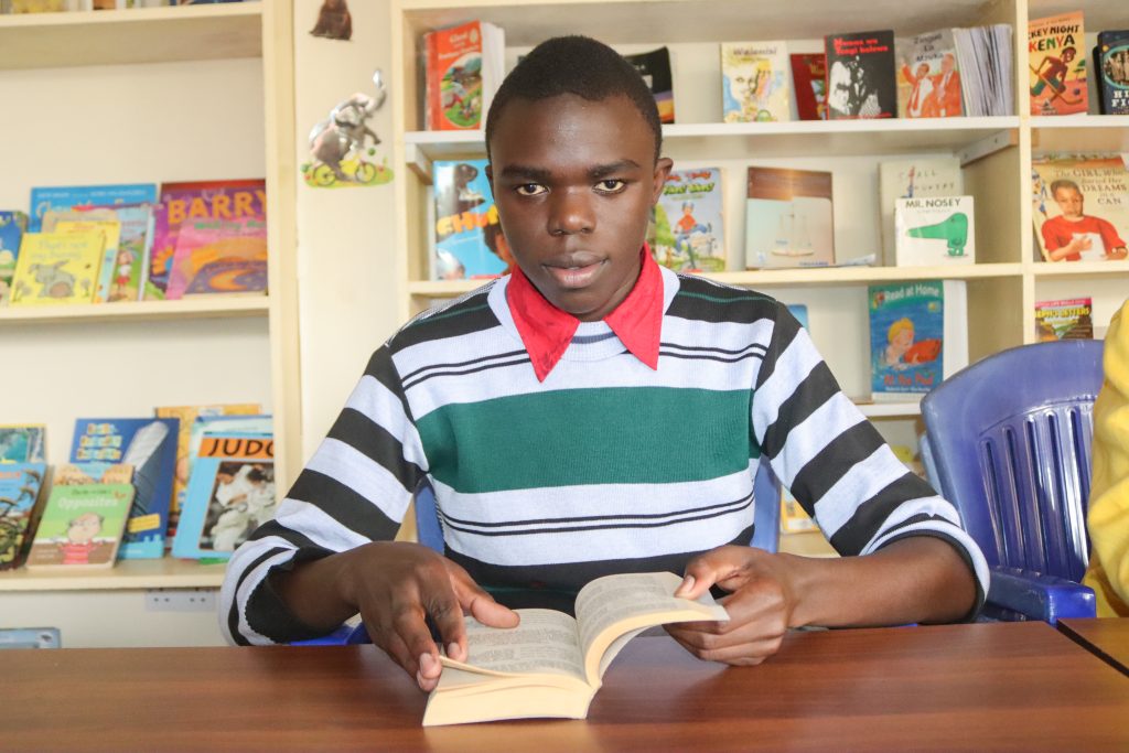 A photograph captures Rolex, a young student with an air of focus and eagerness, engrossed in a captivating novel.